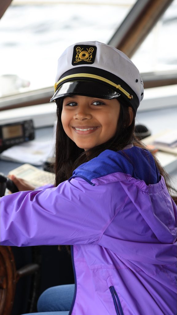 Young girl smiling, wearing captain's hat and purple jacket on a Killarney Lake Tour boat, holding the wheel.