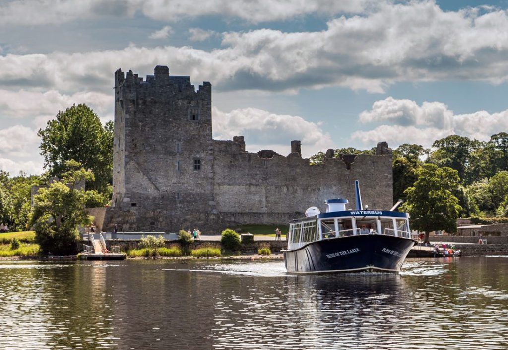 Boat tour on Killarney Lake with historic castle backdrop on a sunny day and lush greenery