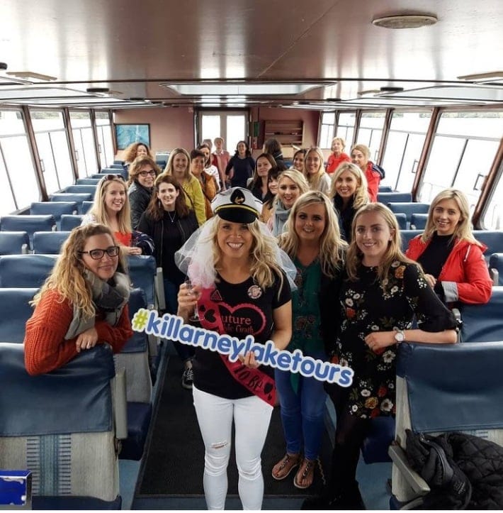 A group on a Killarney Lake Tours boat, the focus is a bride-to-be holding a #killarneylaketours sign.