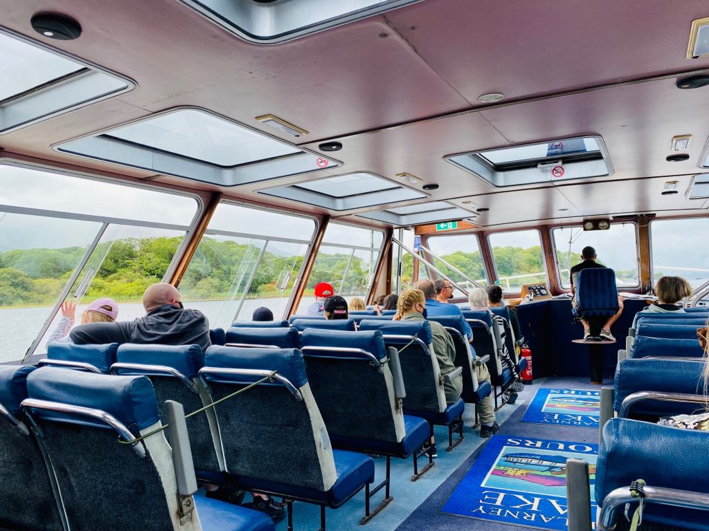 Passengers enjoy scenic views from a boat on Killarney Lake Tour, with lush greenery visible through windows.