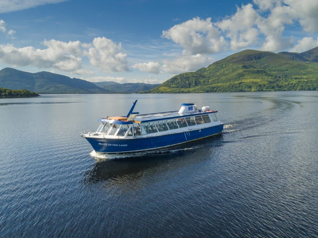 Tour boat on Killarney Lake with lush green mountains in the background under a blue sky.