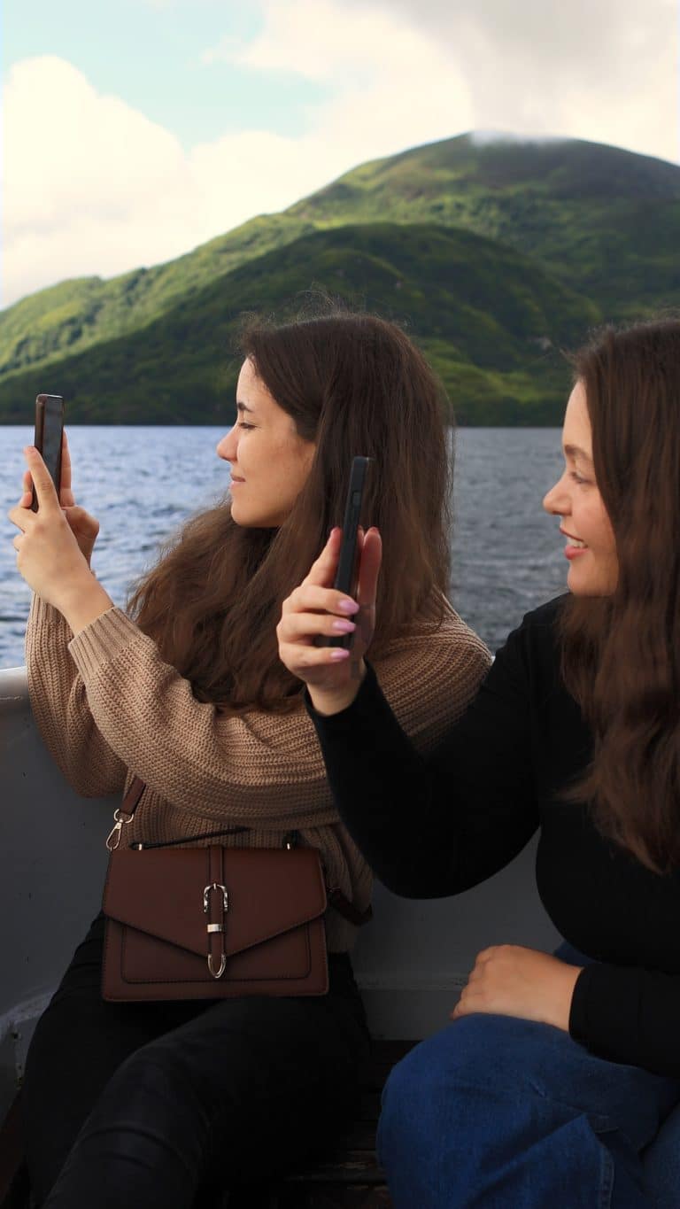 Two women on a boat tour, taking photos of a lush green hill across Killarney Lake.