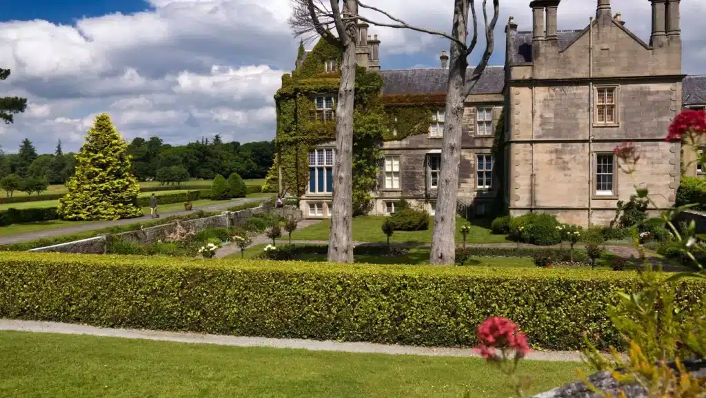 Muckross House with ivy-covered walls, nestled in gardens near Killarney, under a partly cloudy sky.